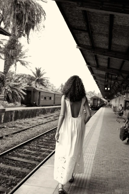 Monochrome shot of a woman in a dress walking along train tracks with palm trees in the background at an outdoor train station.