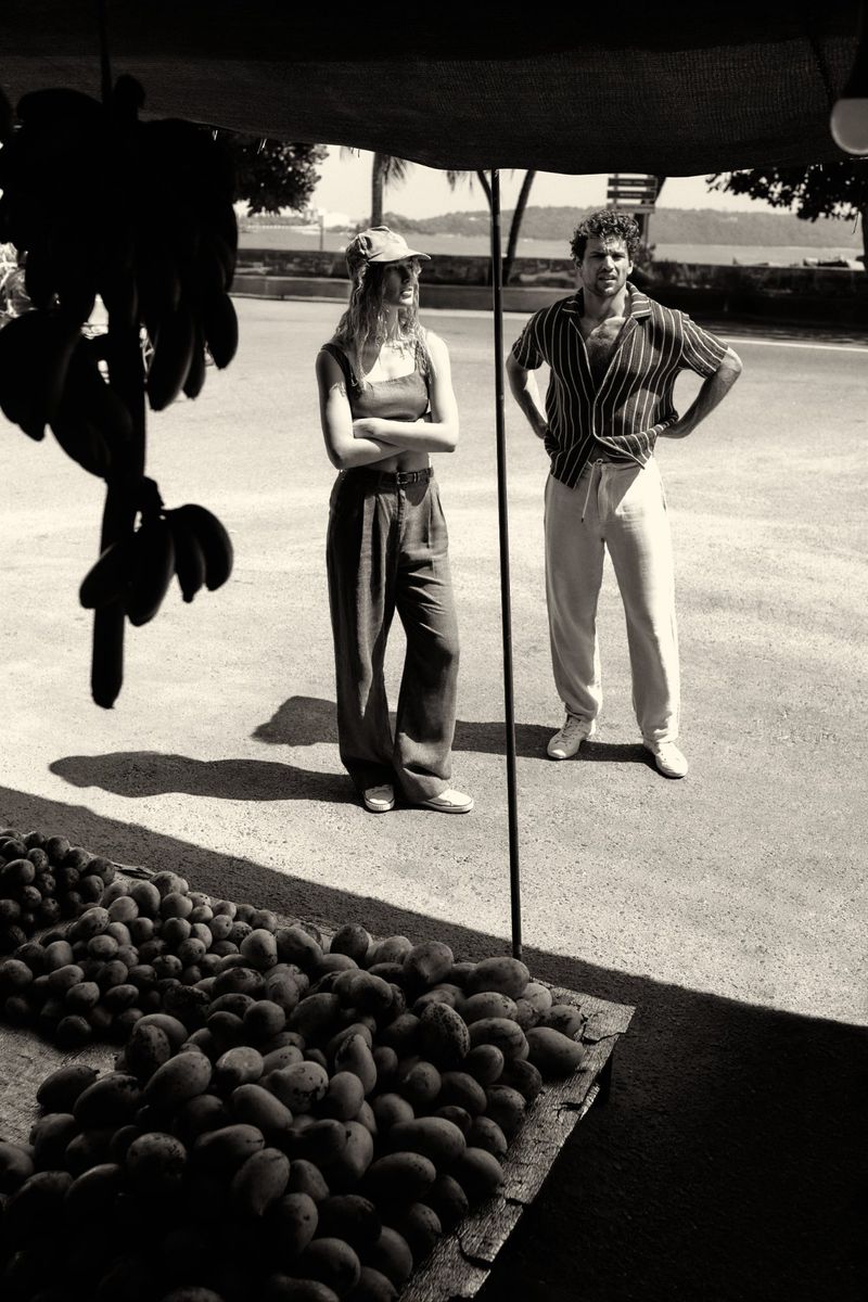 Black and white shot of a woman in a cap and a man in a striped shirt standing next to a table of fruit under an awning.