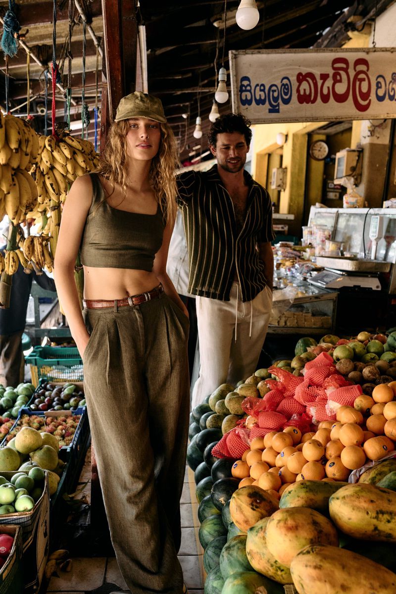 A stylish woman poses at a market, wearing an olive-green crop top and trousers. A man stands in the background among colorful fruits.