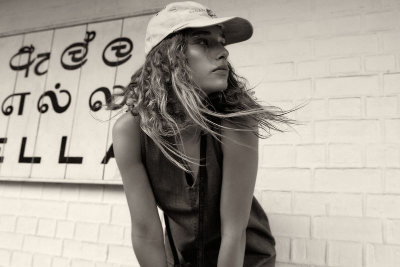 A woman with wavy hair and a cap poses in front of a textured wall with signage in black and white, evoking a casual fashion editorial.