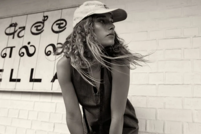 A woman with wavy hair and a cap poses in front of a textured wall with signage in black and white, evoking a casual fashion editorial.