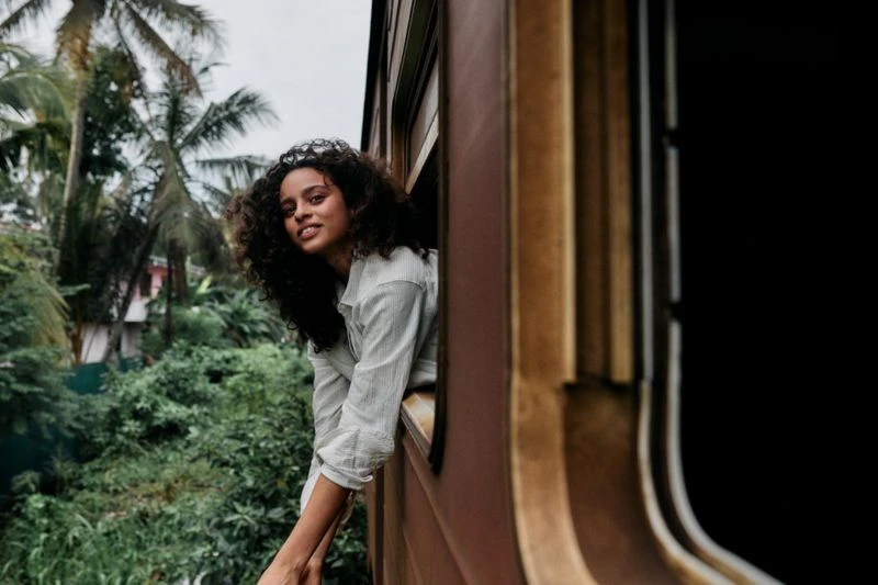 Young woman with curly hair leans out of a train window against a backdrop of lush green vegetation, enjoying a scenic view.