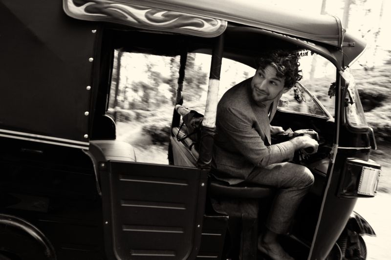 Monochrome shot of a curly-haired man driving an auto rickshaw through a road lined with trees, looking back with a smile.