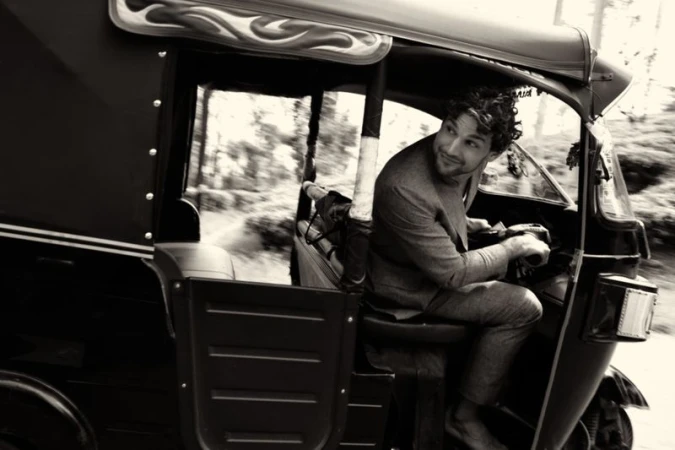 Monochrome shot of a curly-haired man driving an auto rickshaw through a road lined with trees, looking back with a smile.