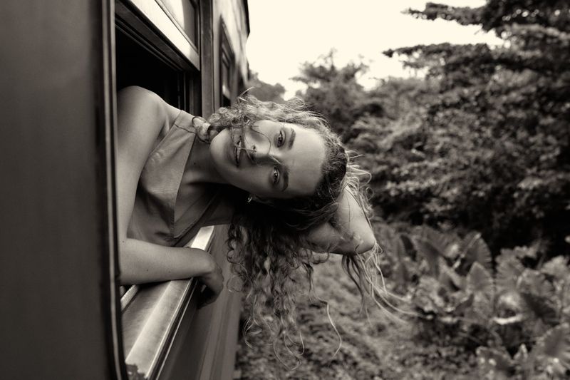 Monochromatic portrait of a curly-haired woman leaning out a train window, with green foliage visible in the background during daytime.