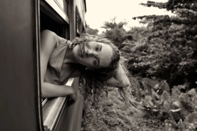 Monochromatic portrait of a curly-haired woman leaning out a train window, with green foliage visible in the background during daytime.