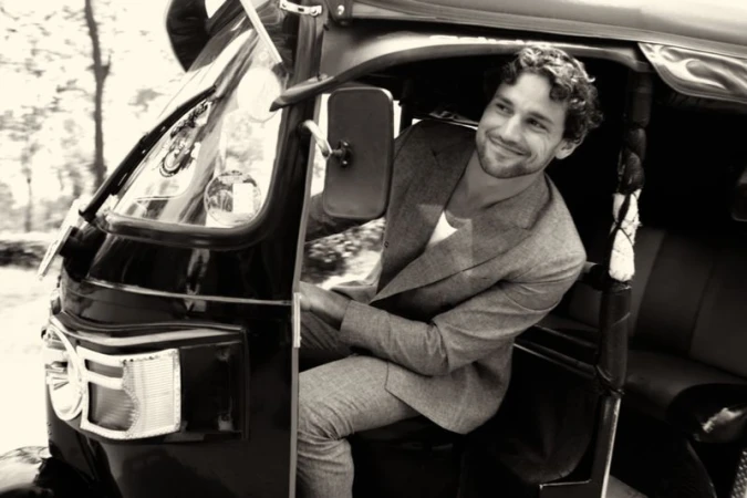Monochrome portrait of a man inside an auto rickshaw, smiling and looking out, wearing a gray suit jacket with blurred trees in the background.