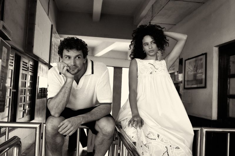 Black and white shot of a man in a polo shirt and a woman in a white dress sitting on metal railings.