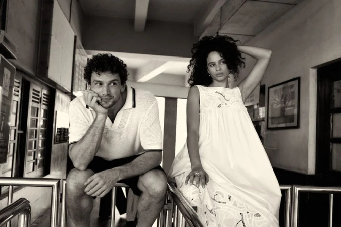 Black and white shot of a man in a polo shirt and a woman in a white dress sitting on metal railings.