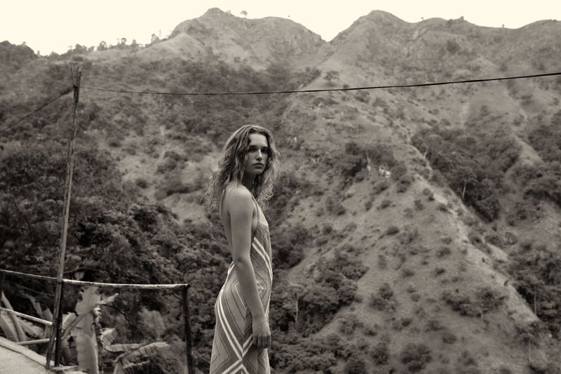 Woman in dress stands in front of mountainous landscape with lush greenery in monochrome image, creating a natural, scenic view.