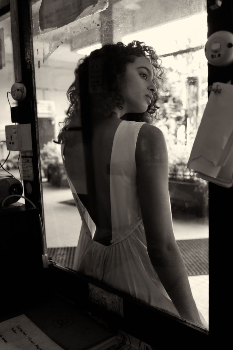 Monochrome portrait of a young woman with curly hair in a low-back dress looking out a window with natural lighting, indoors.