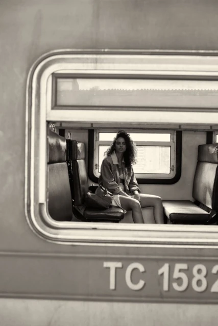 A woman with curly hair sits inside an old train car near the window, wearing a jacket and dress, captured in desaturated tones.