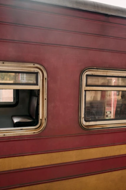 Close-up of the exterior of a maroon and yellow train car featuring two windows, one with an interior seat visible.