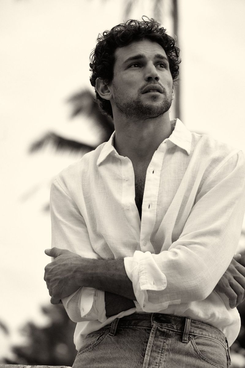 Sepia portrait of a curly-haired man in a white linen shirt and blue jeans looking up with crossed arms against a sky and palm trees.