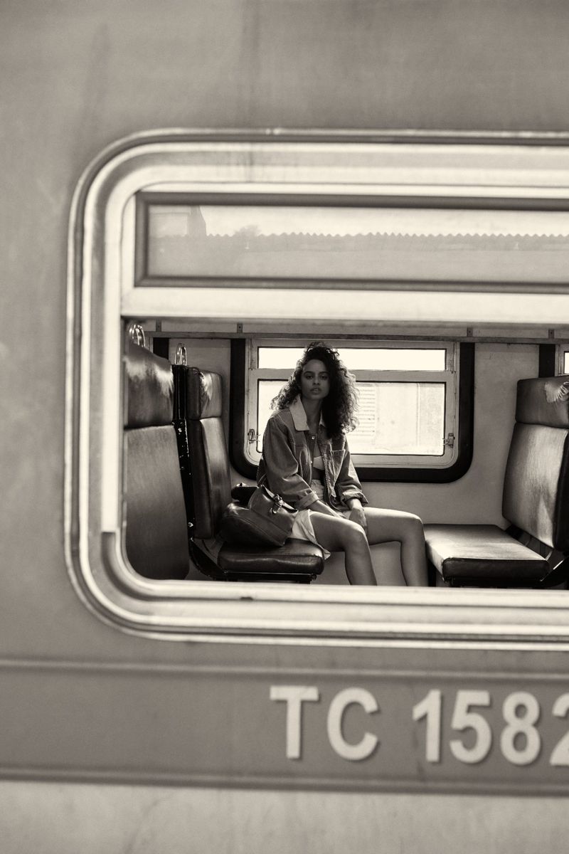 Monochrome shot of a curly-haired woman sitting inside a vintage train car, wearing a denim jacket and light dress, looking out the window.