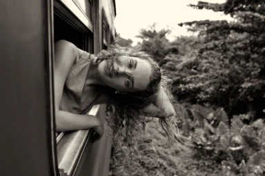 Woman with curly hair leaning out of a moving train window looking backward into lush green foliage