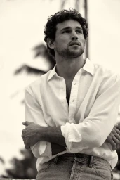 Man with curly hair wearing a white collared shirt and jeans posing outdoors with palm trees in soft focus background.
