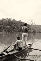 Monochrome image of two models on a wooden boat in a lake with a wooded shore background.