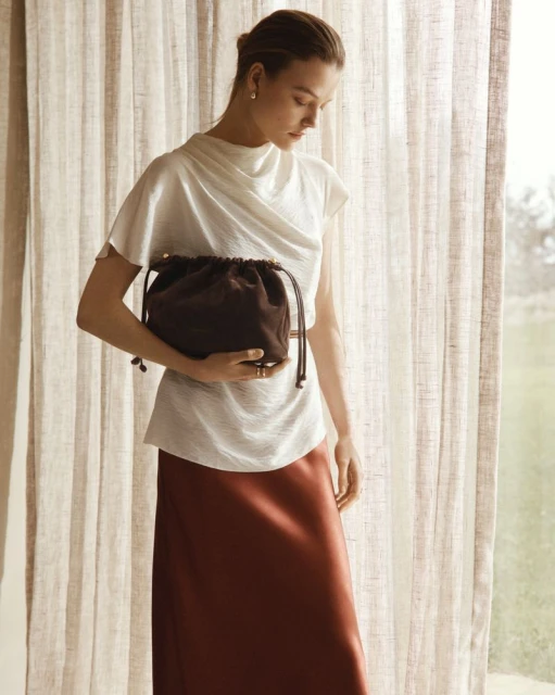 Elegant woman in white top and rust-colored skirt holding a dark brown drawstring bag, posing in front of sheer curtains.