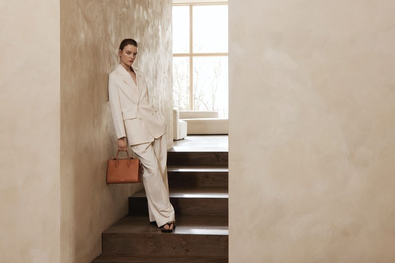 Elegant woman in cream suit and black sandals poses on stairs with a brown handbag, set against a minimalist cream textured wall.
