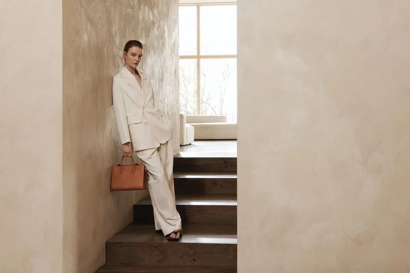 Elegant woman in cream suit and black sandals poses on stairs with a brown handbag, set against a minimalist cream textured wall.