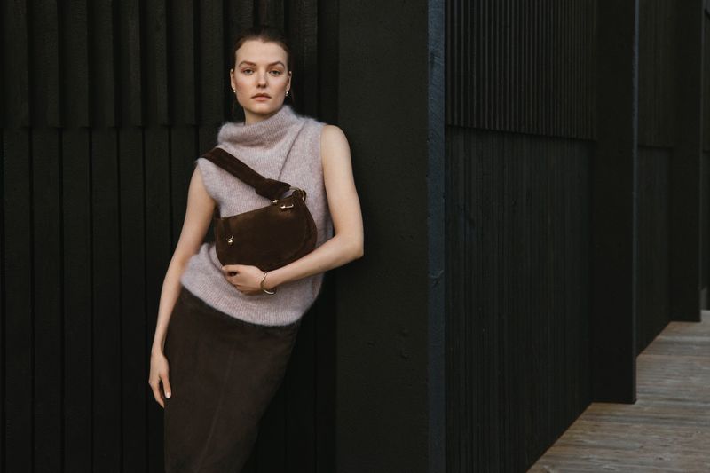 A woman with a brown purse poses against a dark wood wall, wearing a mauve sweater and skirt, illuminated by soft studio lighting.
