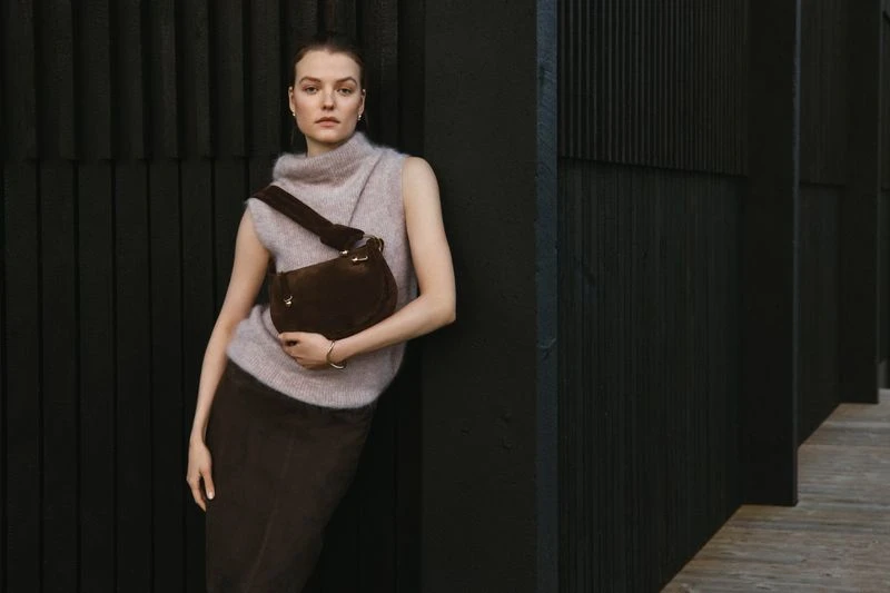 A woman with a brown purse poses against a dark wood wall, wearing a mauve sweater and skirt, illuminated by soft studio lighting.