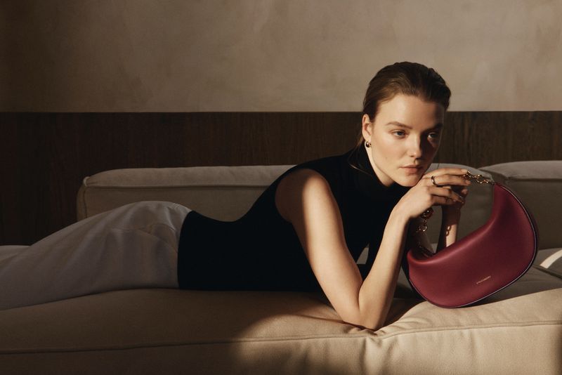 Brunette woman in a black sleeveless top reclines on a cream couch indoors with a burgundy purse featuring a gold chain.