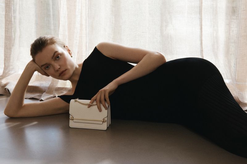Model reclining on floor in a black dress holding a white structured handbag against linen curtains