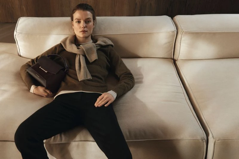 A woman in a sweater and dark pants reclines on a cream couch with a dark handbag, creating a stylish, relaxed indoor fashion shot.