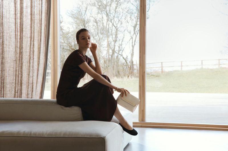 Model Roos Abels seated on a light sofa holding a white handbag in a bright interior setting by Amie Milne