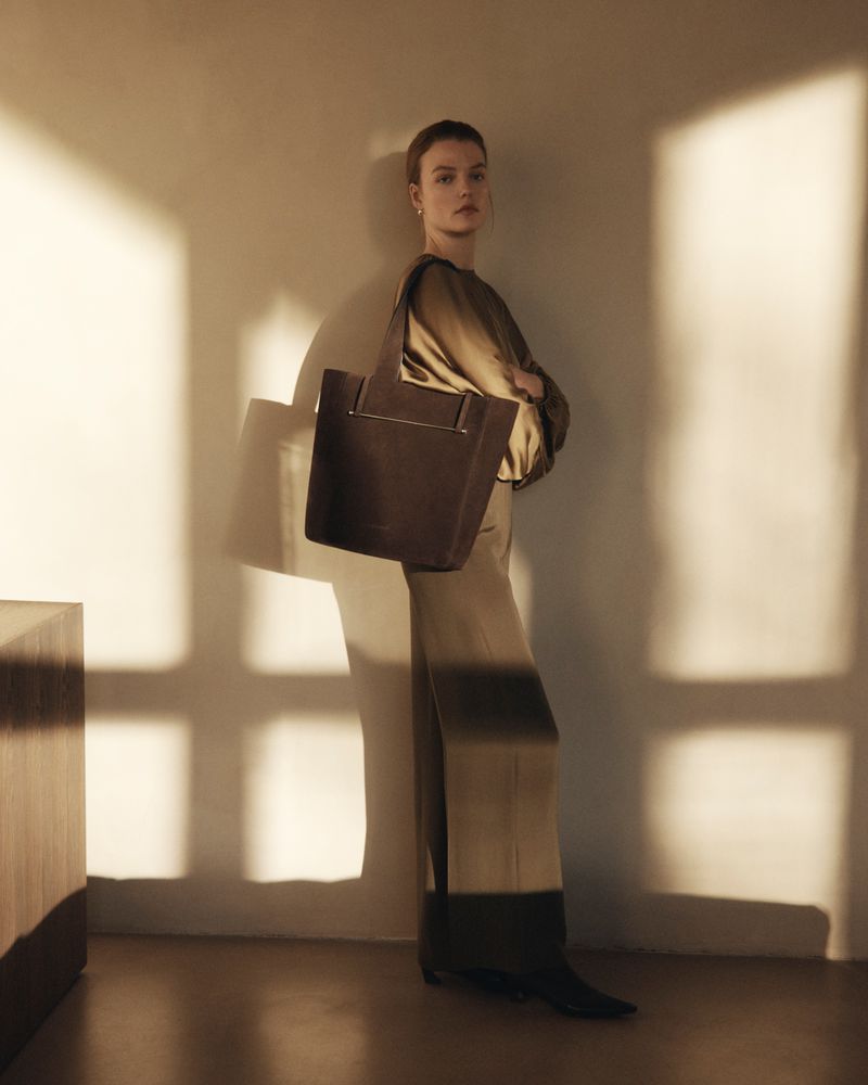 Model wearing a silky olive top and skirt holding a large dark tote bag in warm sunlight with window shadows.