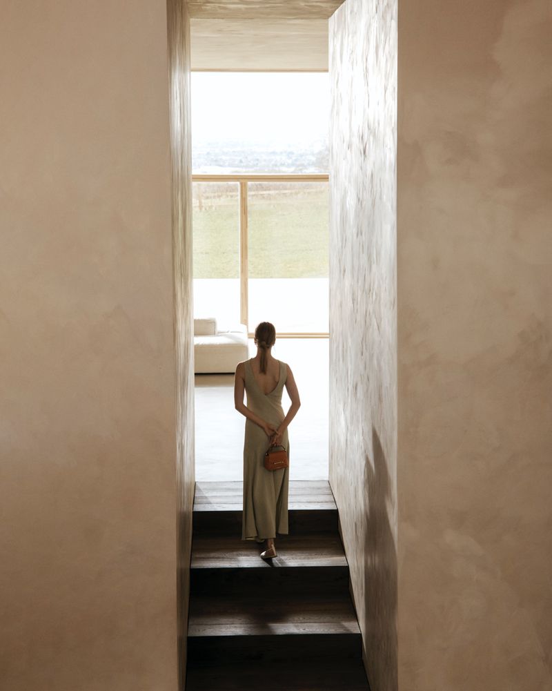 Woman walks down stairs in light-filled minimalist space, wearing a dress and carrying a small purse; interior with a view to the outside.