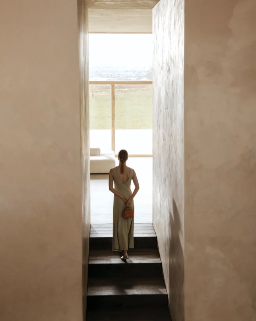 Woman walks down stairs in light-filled minimalist space, wearing a dress and carrying a small purse; interior with a view to the outside.