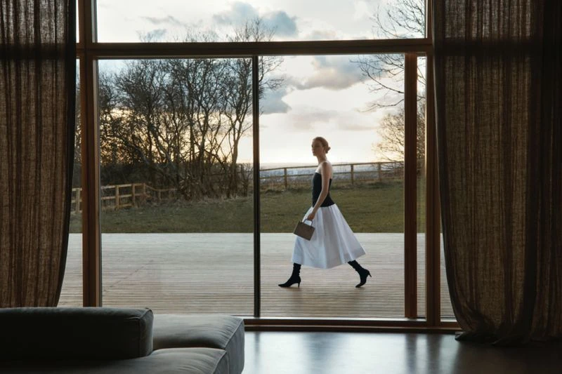 An adult woman in a black top and white skirt walks in a minimalist home interior with a view of a green field through large windows.