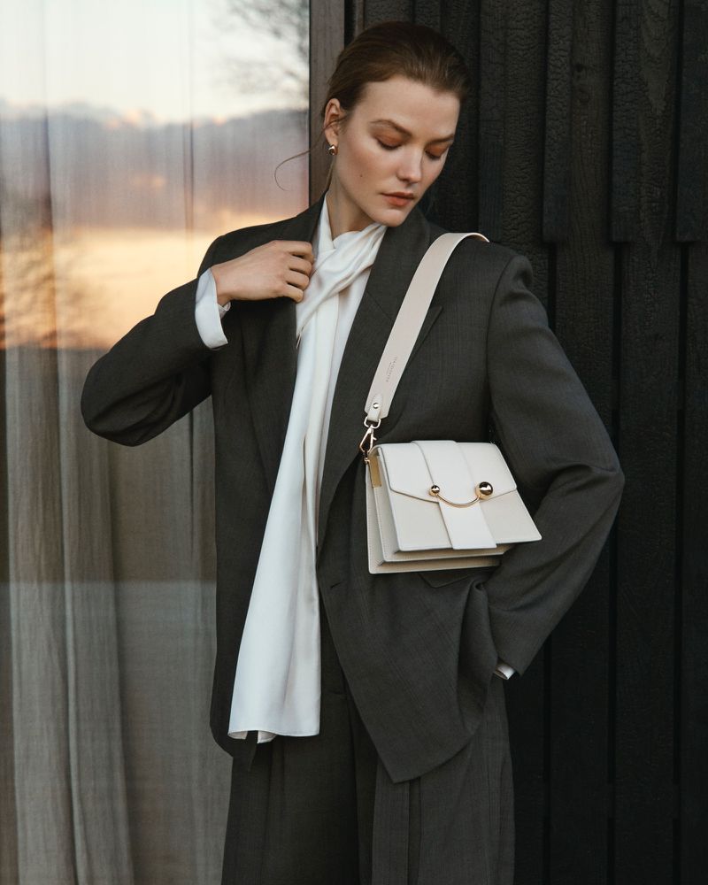 Woman in dark grey suit and white scarf with a white cross-body bag, posing against a dark wood wall with a neutral expression.