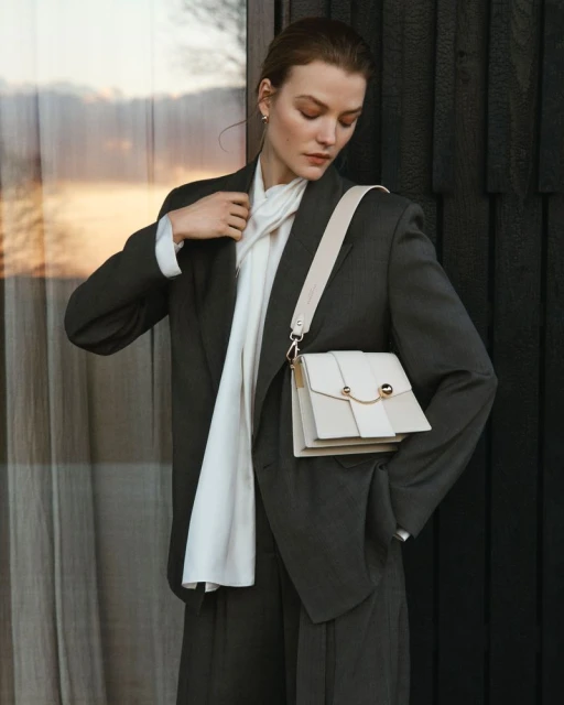 Woman in dark grey suit and white scarf with a white cross-body bag, posing against a dark wood wall with a neutral expression.