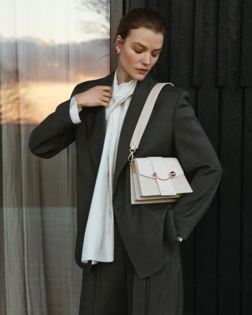 Woman in dark grey suit and white scarf with a white cross-body bag, posing against a dark wood wall with a neutral expression.