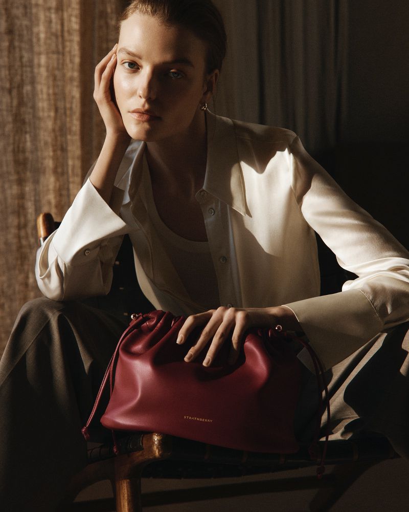 Fashionable adult woman poses indoors with a maroon Strathberry handbag, wearing a white blouse and grey trousers, lit by soft natural light.