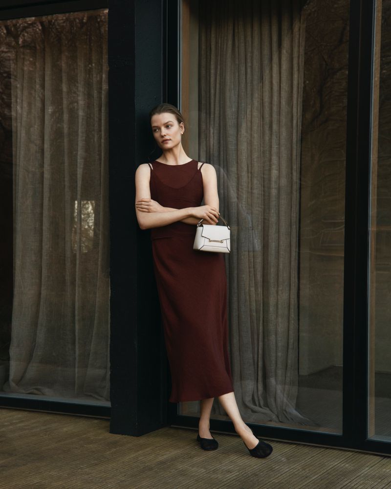 Indoor portrait of a woman in a sleeveless maroon dress, holding a structured cream-colored handbag, standing near large windows.