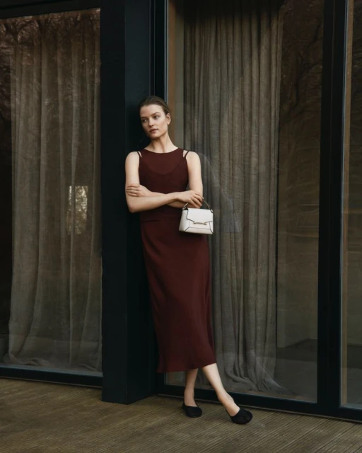 Indoor portrait of a woman in a sleeveless maroon dress, holding a structured cream-colored handbag, standing near large windows.