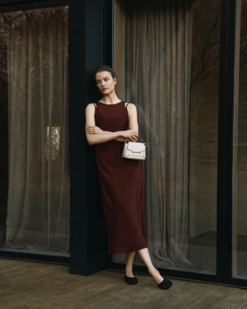 Indoor portrait of a woman in a sleeveless maroon dress, holding a structured cream-colored handbag, standing near large windows.
