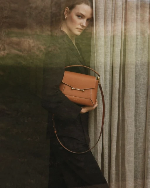 Adult woman in black suit jacket poses with brown leather handbag next to a curtain. Fashion editorial photography.