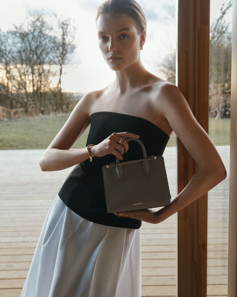Woman in black top and white skirt holds a gray handbag, posing in an indoor setting with trees visible through a window.