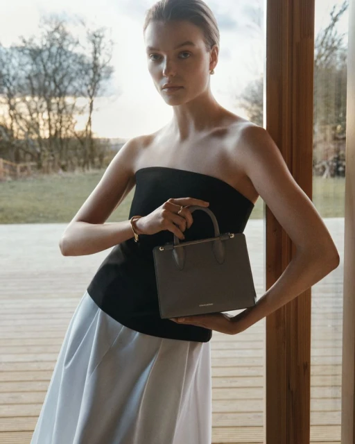 Woman in black top and white skirt holds a gray handbag, posing in an indoor setting with trees visible through a window.