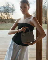 Model Roos Abels holding a grey handbag while leaning against a wooden doorframe in a countryside setting