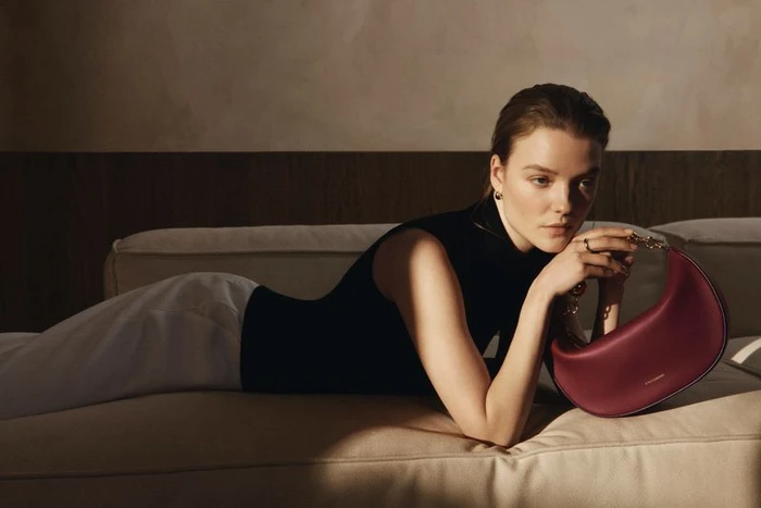 Model reclining on a cream sofa holding a burgundy leather handbag wearing a black sleeveless top and white trousers