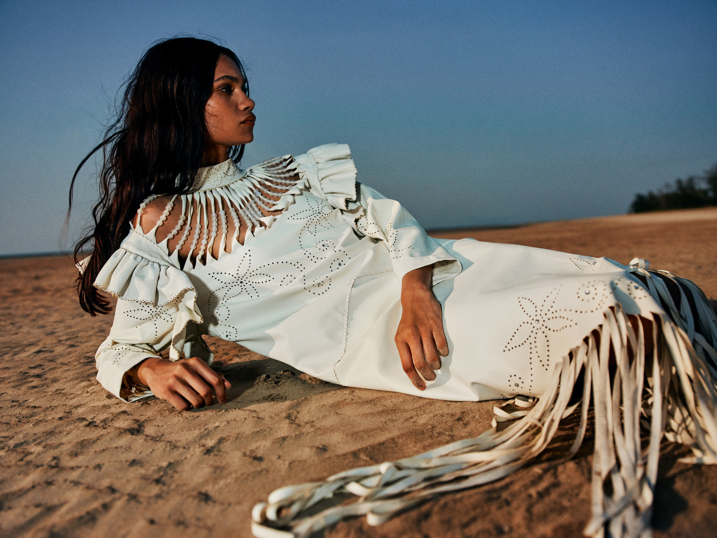 A woman with dark hair lies on a sandy beach in a white, embellished dress with fringe, under a clear blue sky during daylight hours.
