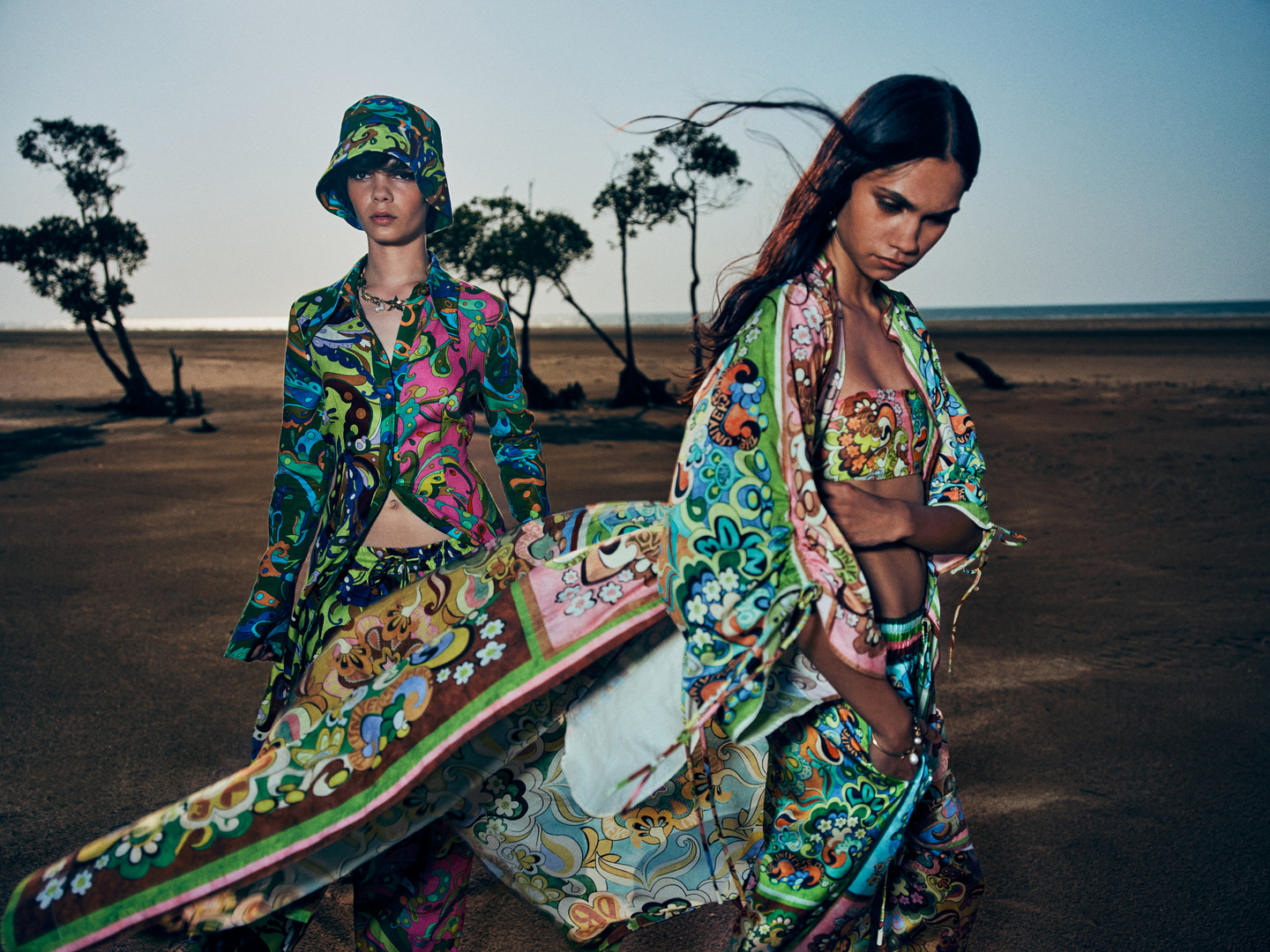 Two female fashion models wearing matching patterned outfits pose on a sandy beach with sparse trees under a light sky.