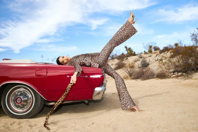 An adult female in a leopard-print suit poses on a red convertible car under a blue sky with desert vegetation in the background.